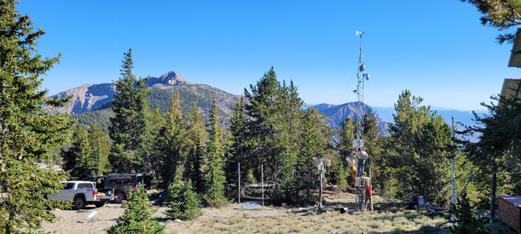View from the top of a mountain where a DRI truck is parked and researchers are working on a weather station and surrounded by pine trees.