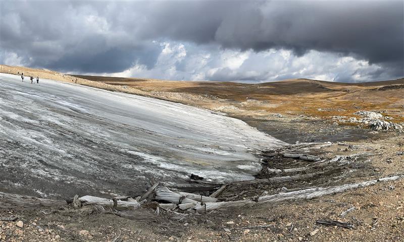 Landscape that includes people standing on a large ice sheet with hills and dark clouds in the distance.