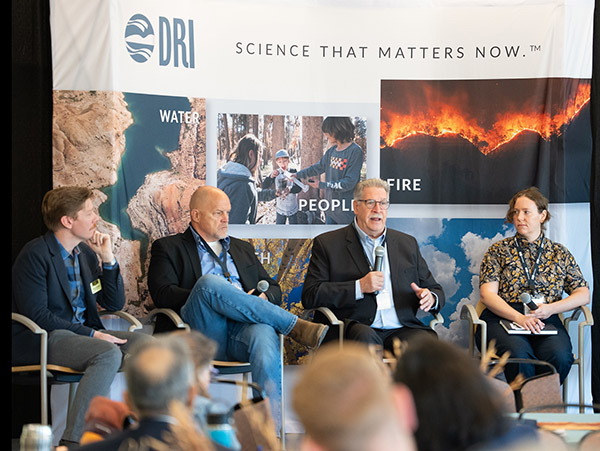 A panel of 4 speakers sits on chairs in front of a DRI banner holding microphones. 