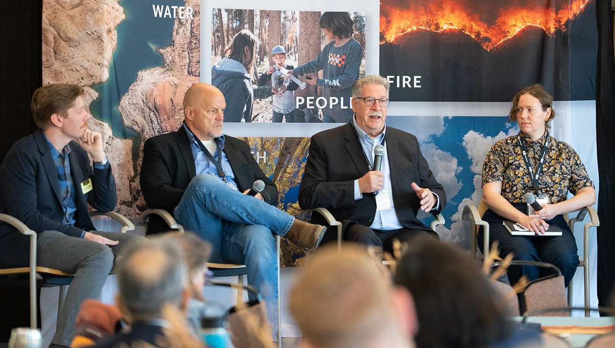 A panel of 4 speakers sits on chairs in front of a DRI banner holding microphones.