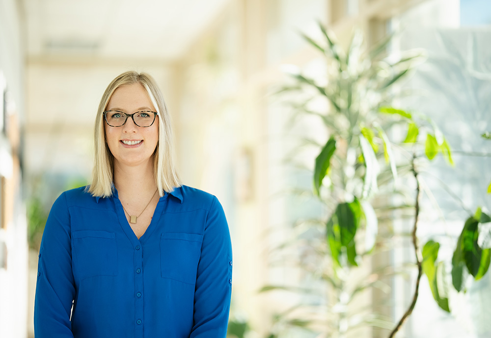 Alexa Daniels headshot in a bright hallway with plants wearing a blue blouse and glasses.
