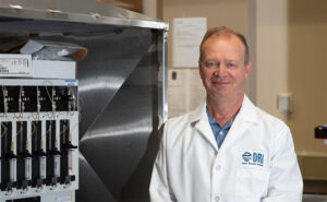 Researcher stands by lab equipment in white lab coat with DRI above the pocket.