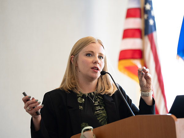 Ashley presenting at a podium with a microphone and American flag in the background.