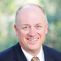 Bruce Yerman smiling for the camera in a suit and tie outside with trees in the background.