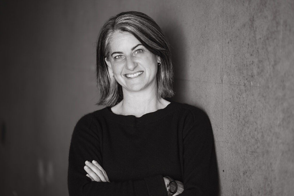 Black and white headshot of Cayenne leaning against a wall with arms folded and smiling.