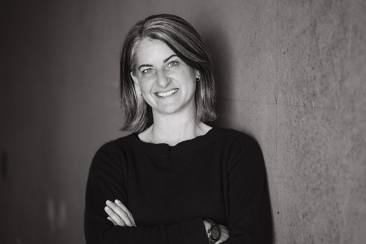 Cayenne Engel Featured Image Black and white headshot of Cayenne leaning against a wall with arms folded and smiling.