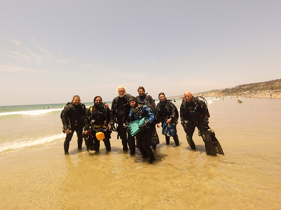 Seven divers stand in a group in wet suits smiling for the camera on a beach.