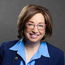 Headshot of Naomi Duerr wearing red glasses and a dark blue suit against a neutral background. 