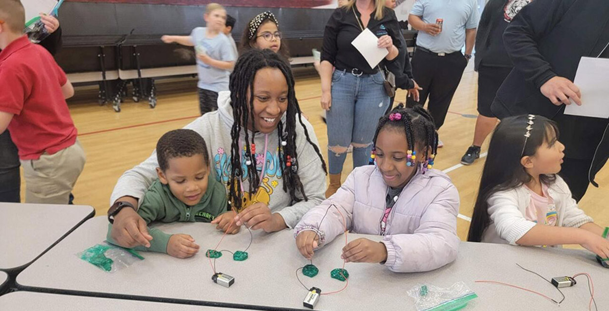 An adult and two children are working on building circuits at a table at a community event.
