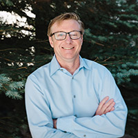 Headshot of Eric Wilcox outside in front of pine trees wearing glasses and a blue collared shirt. 