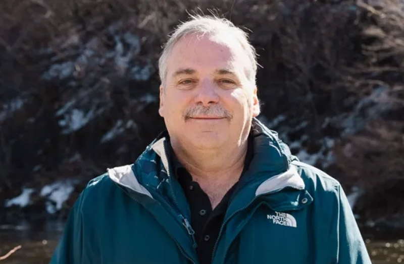 Frank McDonough wearing a jacket and standing in front a stream with the sun shining.
