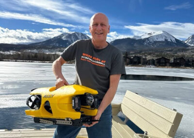 A researcher is standing on a dock holding a remote operated vehicle (ROV) in front of a frozen body of water.