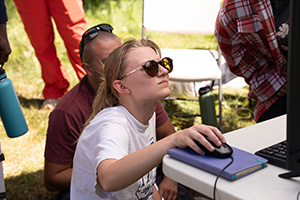 A student looks up at a computer screen while controlling the mouse out in the field on a project with others onlooking. 
