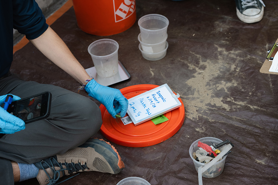 Intern sitting on the ground and documenting items found on the beach with a white board, bucket lid, and phone.