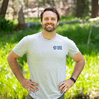 Photo of Justin smiling and standing outside with arms on hips in front of a river bank. 