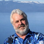Headshot of Lou standing in front of a landscape of a large lake and snowcapped mountains. 