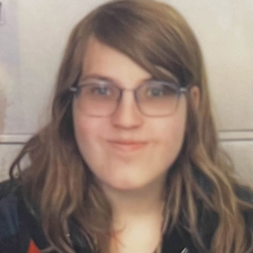 Madeline Veric Group Page Headshot of Madeline smiling and wearing glasses with filing cabinets in the background.