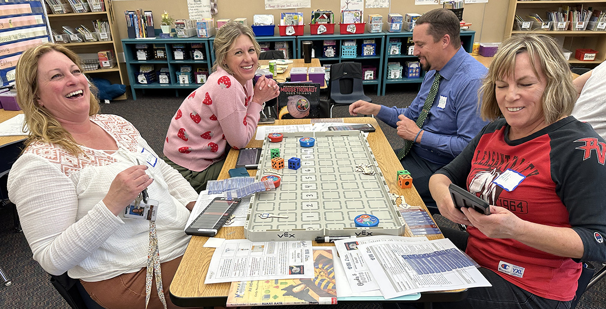 Four teachers smiling and sitting around a rectangular table with a grid and small circular robots. 