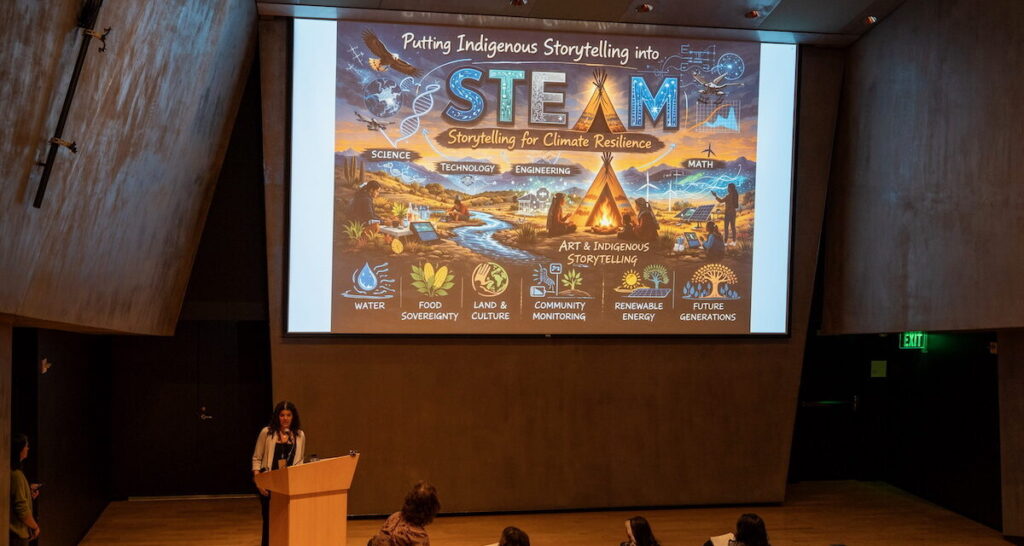 A woman talking on a stage with a projector behind her
