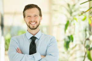 Headshot of Patrick wearing a tie and collared shirt and smiling with arms crossed in a brightly lit hallway full of plants.