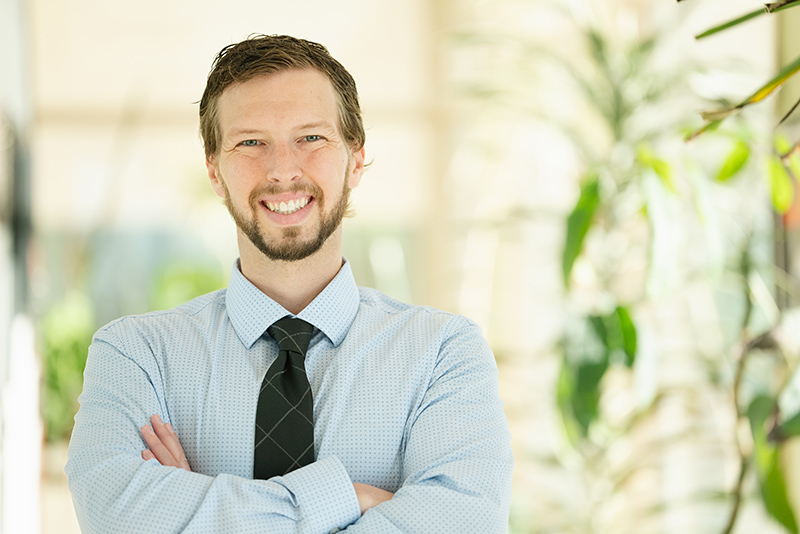 Headshot of Patrick wearing a tie and collared shirt and smiling with arms crossed in a brightly lit hallway full of plants.