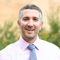 Headshot of Scott outside with greenery in the background and wearing a collared shirt and tie. 