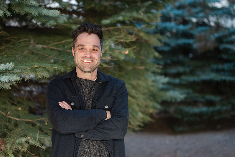 Headshot of Tyler smiling with arms crossed in front of pine trees.