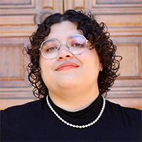Headshot of Vic wearing a black top with pearls and glasses against a patterned background.