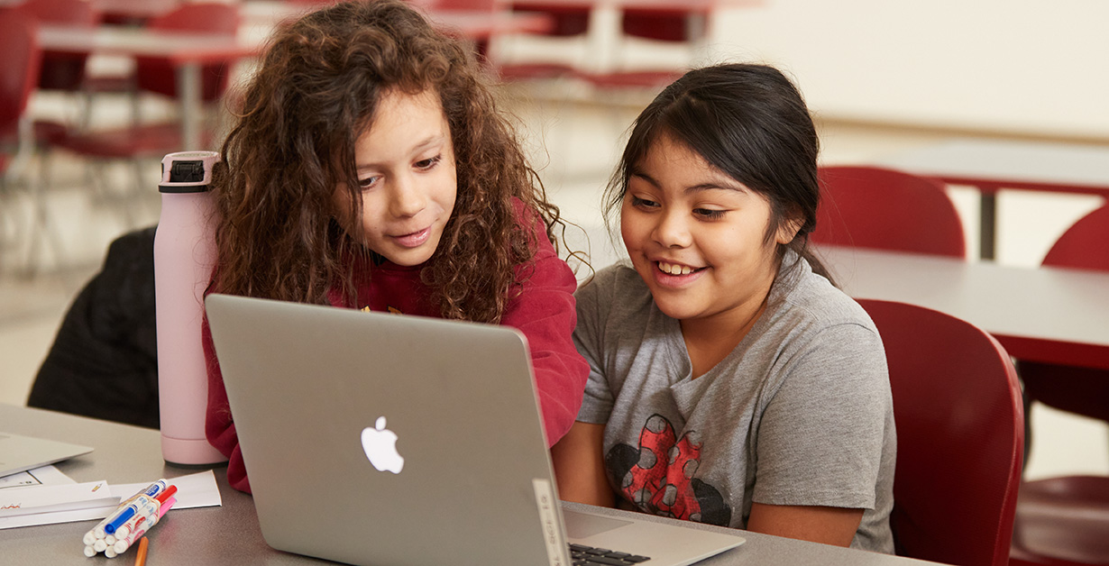 Two students are sitting at a table and working on one laptop and smiling. 