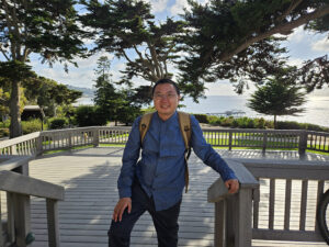 Ziming Ke standing on a deck under trees with the coast of a body of water in the background.
