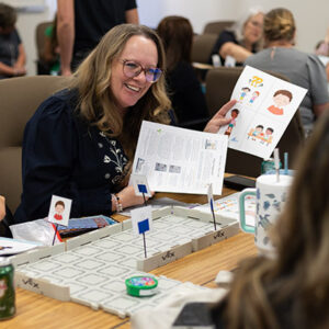 An educator is laughing and holding up two worksheets and looking at the small round robot on the table. 