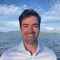 Headshot of Eugene smiling in the sun in front of a body of water. 