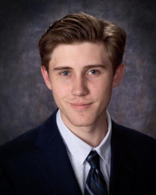 Headshot of Erik Henzl wearing a suit and tie against a dark grey background. 