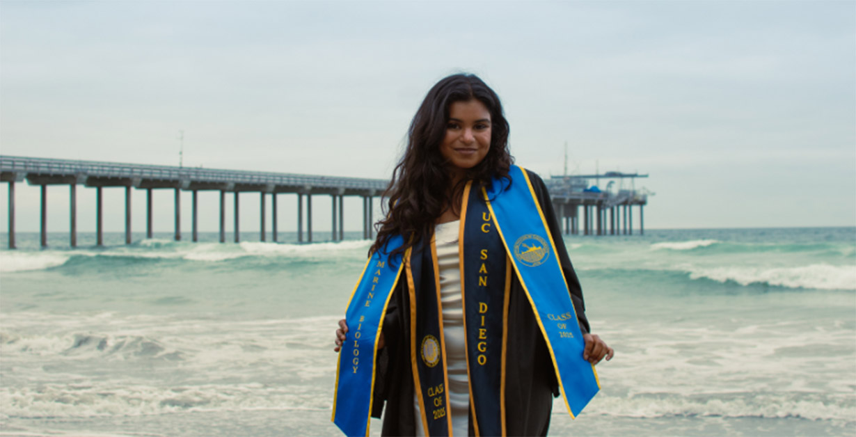 Ria standing on the beach in graduation robes with a pier in the background.