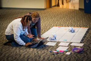 Educators working with a laptop on the floor next to a large map where they are guiding robots on a path.